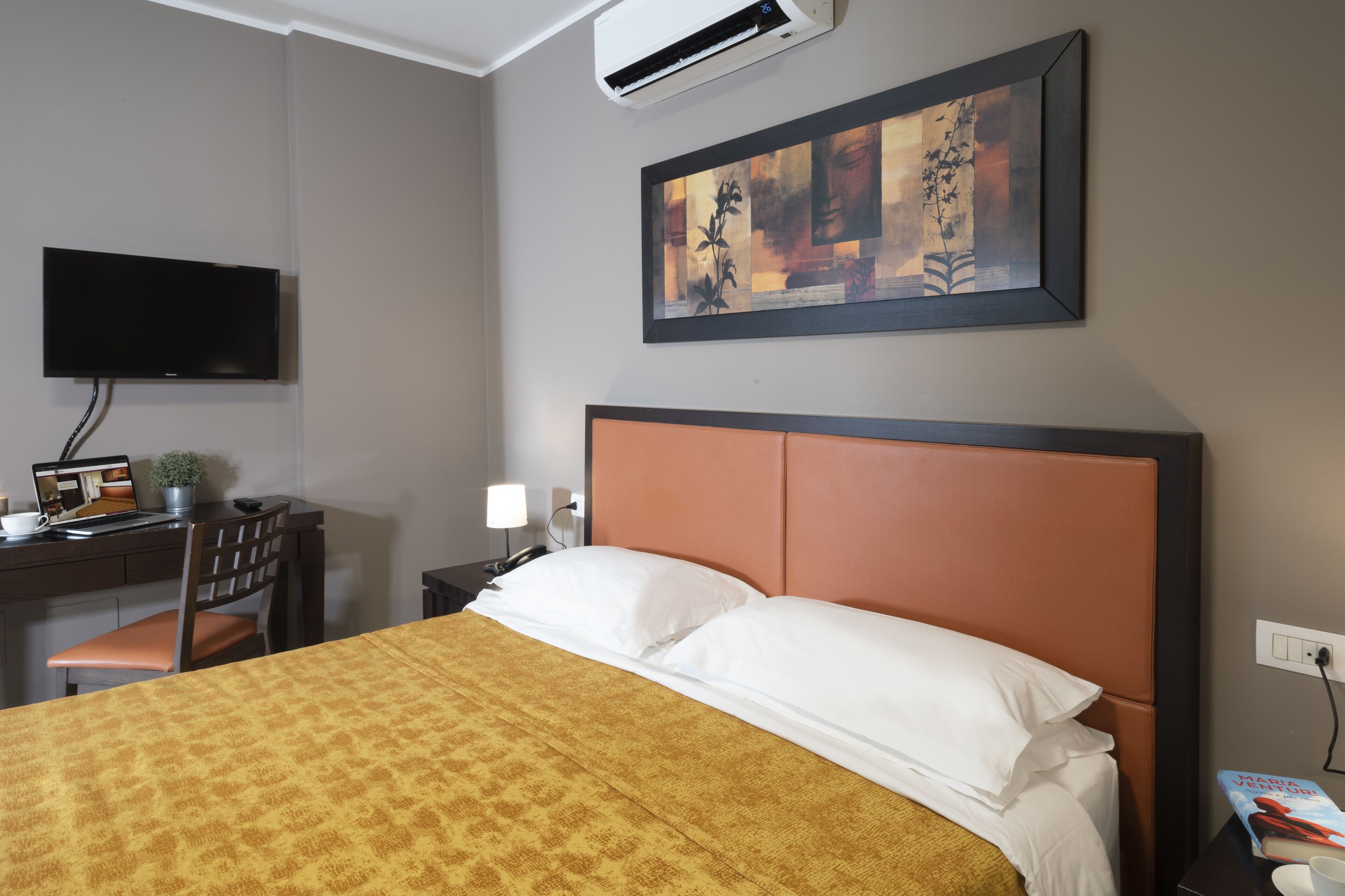 Ein Hotelzimmer mit Bett, Schreibtisch mit Computer, Stuhl, Fernseher und einem Bild an der Wand.