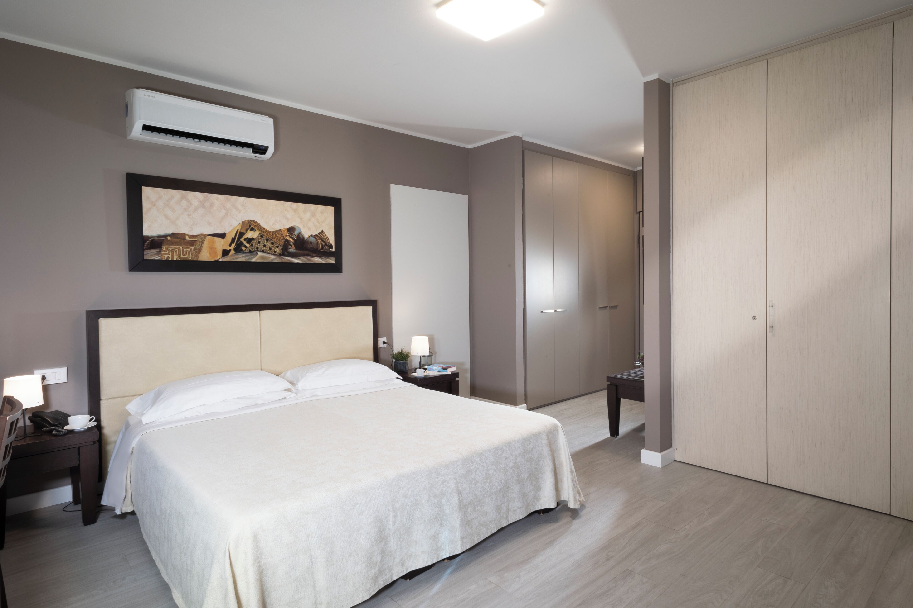 Hotellihuone, jossa on suuri sänky, yöpöytä lampun kanssa, ilmastointilaite, vaatekaappi ja seinällä oleva kehystetty kuva.