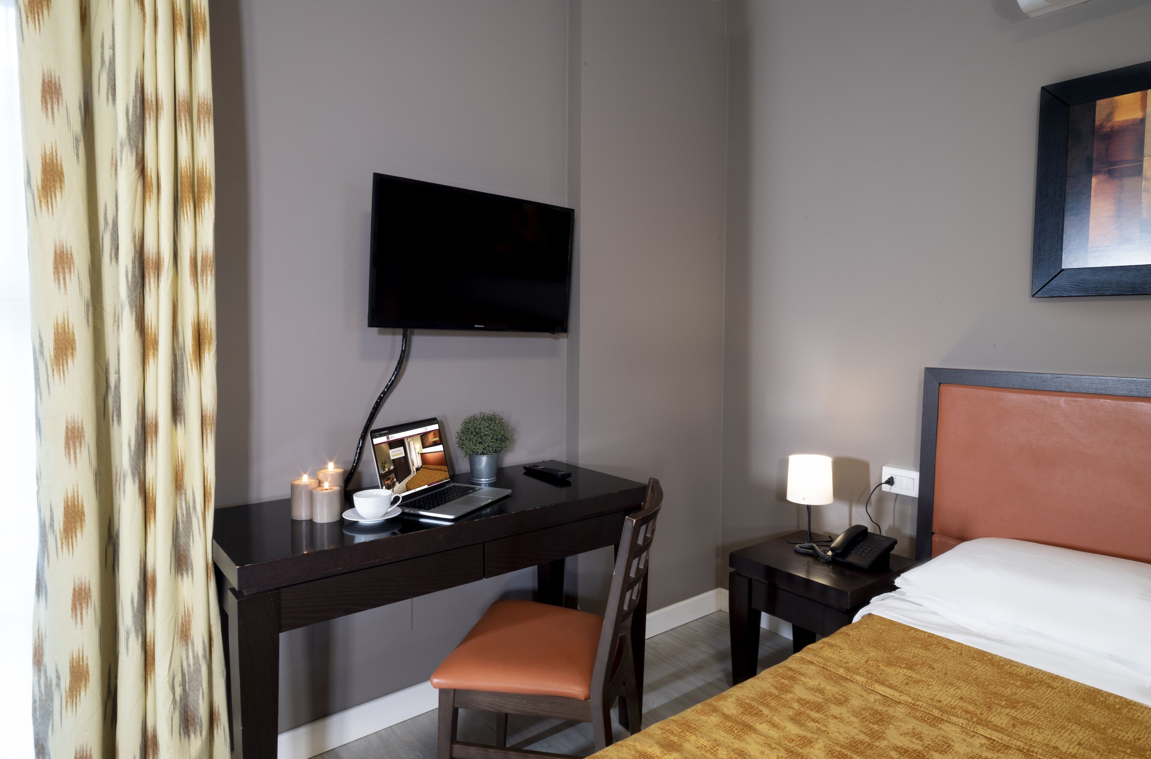 A hotel room with a desk, chair, bed, TV, and bedside table with a lamp.
