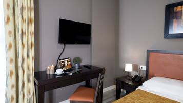 A hotel room with a desk, chair, bed, TV, and bedside table with a lamp.