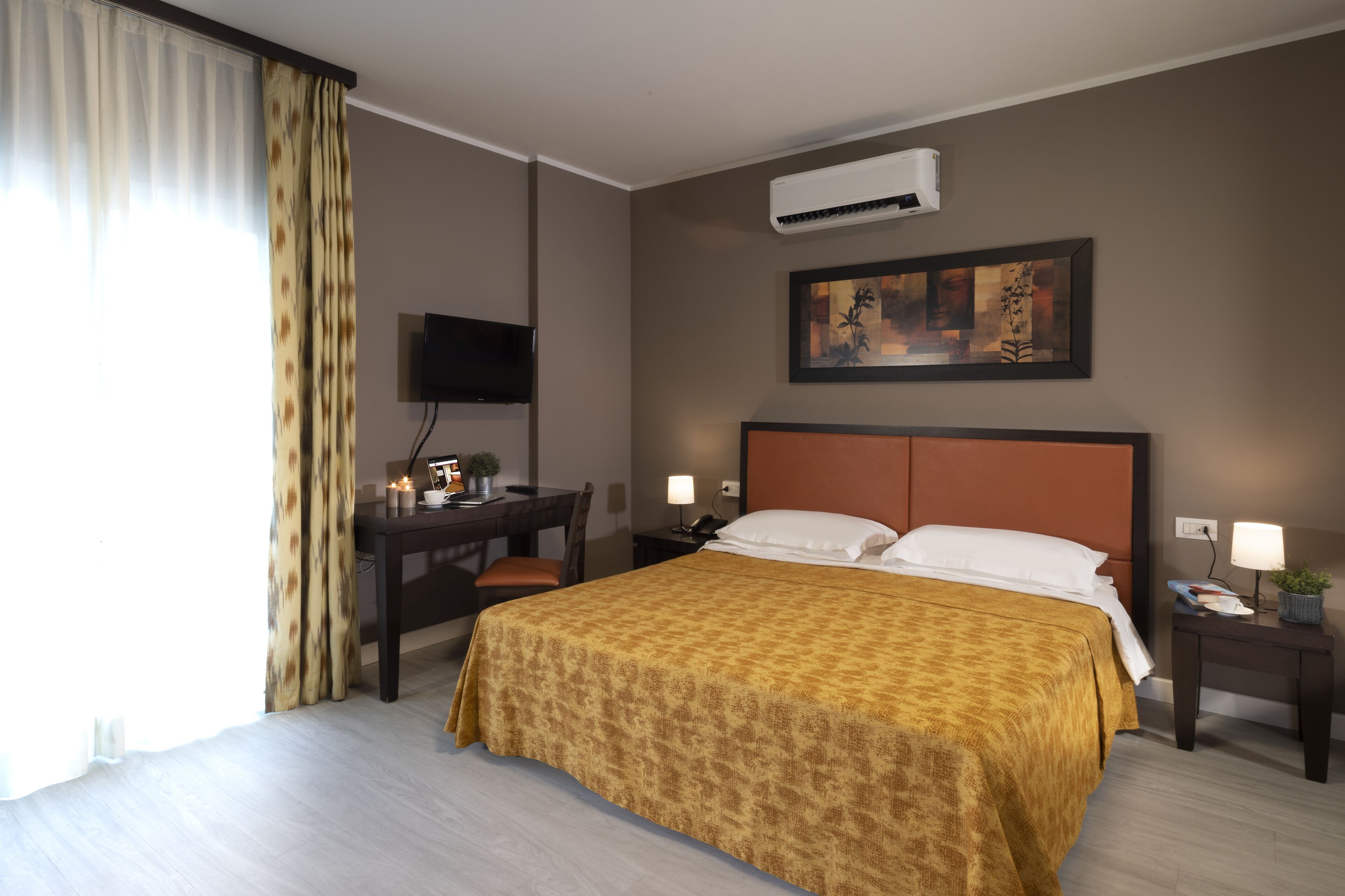 A hotel room with a bed, a desk, a chair, a nightstand, a TV, and a painting on the wall.
