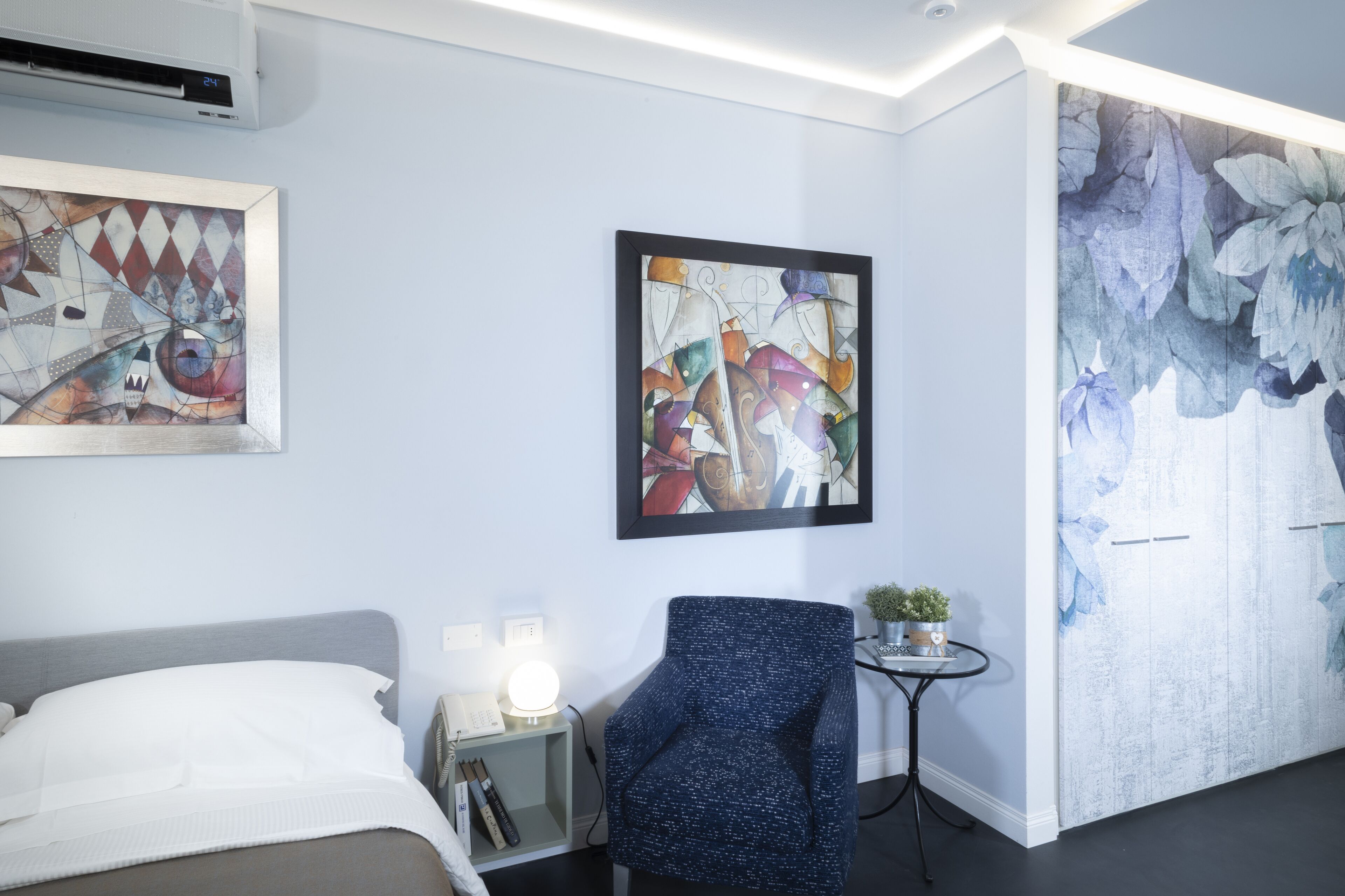 Une chambre d’hôtel avec un lit, une chaise, une petite table avec une lampe, et deux tableaux abstraits encadrés accrochés au mur.