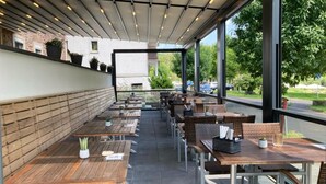 An outdoor cafe with wooden tables and wicker chairs, a covered seating area, and a view of the street and greenery. - Hotel Fritz (Valwig)