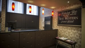 Aula, jossa on tumman puun värinen vastaanottotiski, seinälle kiinnitetty kyltti, jossa lukee: ”Lämpimästi tervetuloa Best Western Grand Hotelliin”, sekä kolme riippuvalaisinta tiskin yläpuolella.