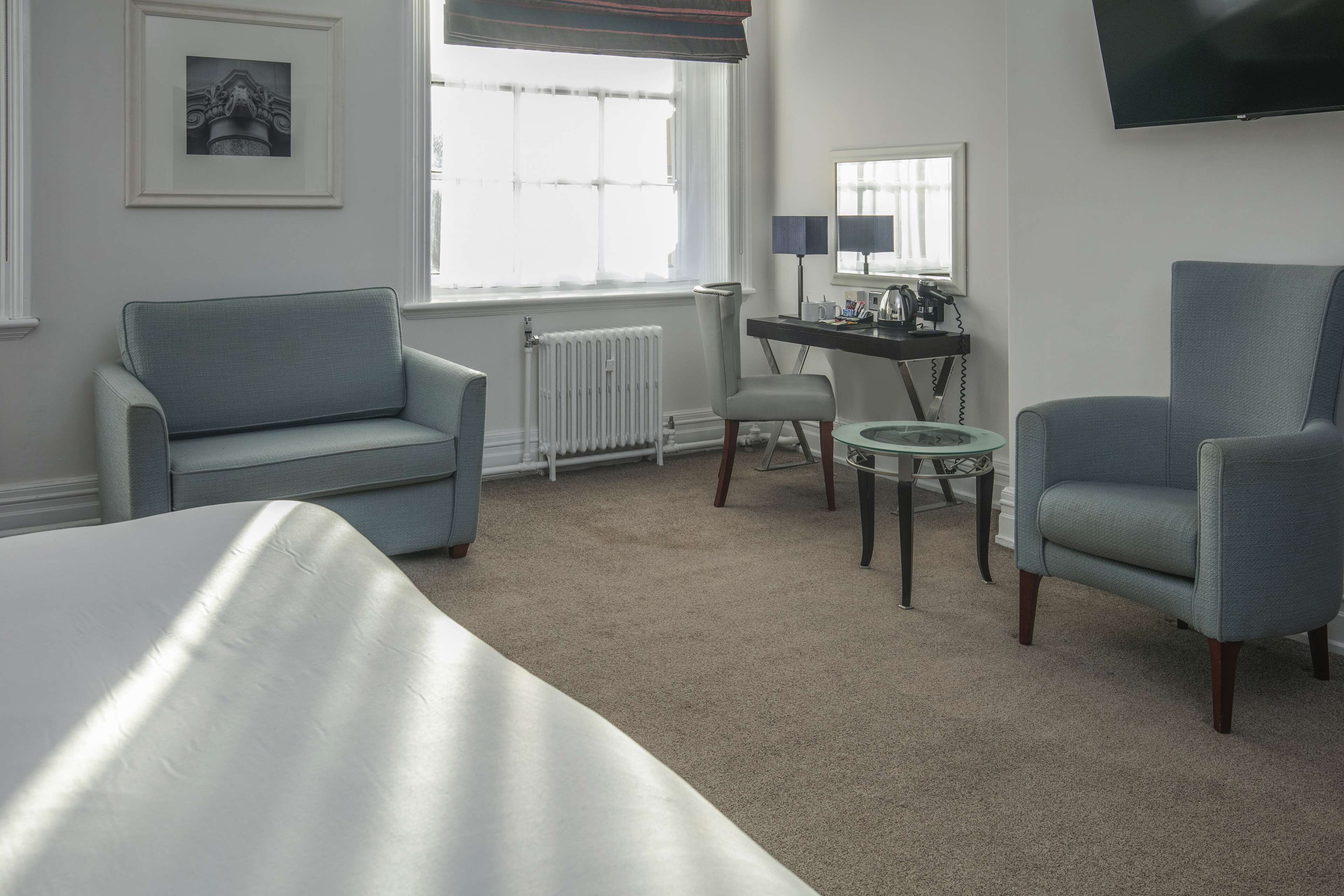 A hotel room with a bed, a grey sofa, a small round table, a desk with a lamp, a chair, and a television on the wall.