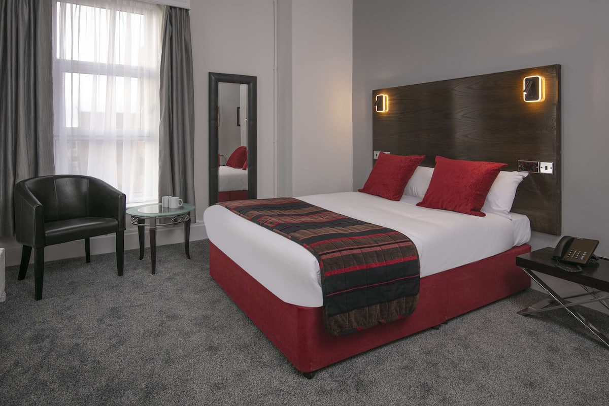 A hotel room with a large bed, red and white bedding, a wooden headboard with a light, a small round table, a chair, and a mirror.