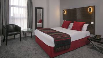 A hotel room with a large bed, red and white bedding, a wooden headboard with a light, a small round table, a chair, and a mirror.