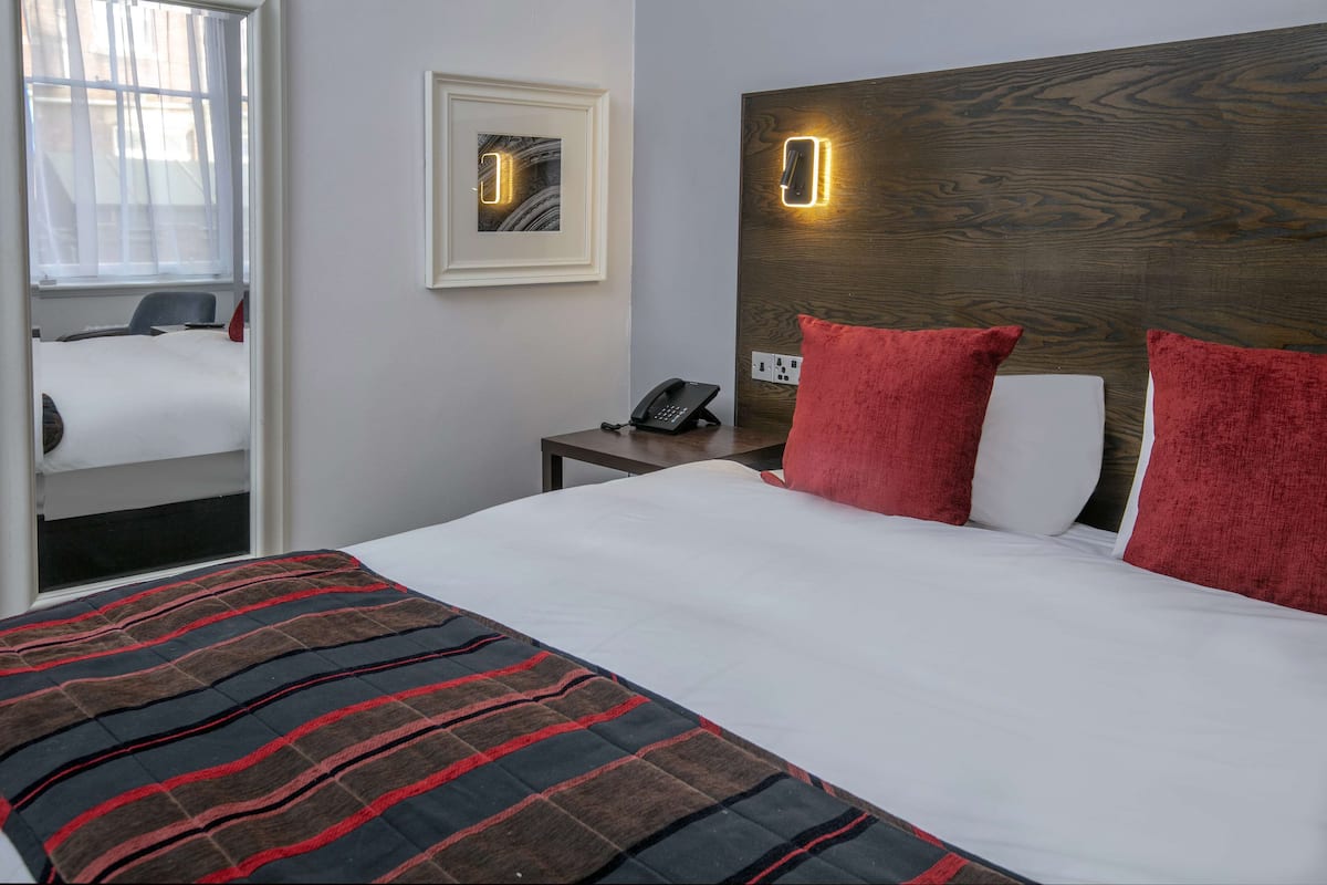 A hotel room with a large bed, a bedside table with a phone, a framed picture on the wall, and a window with sheer curtains.