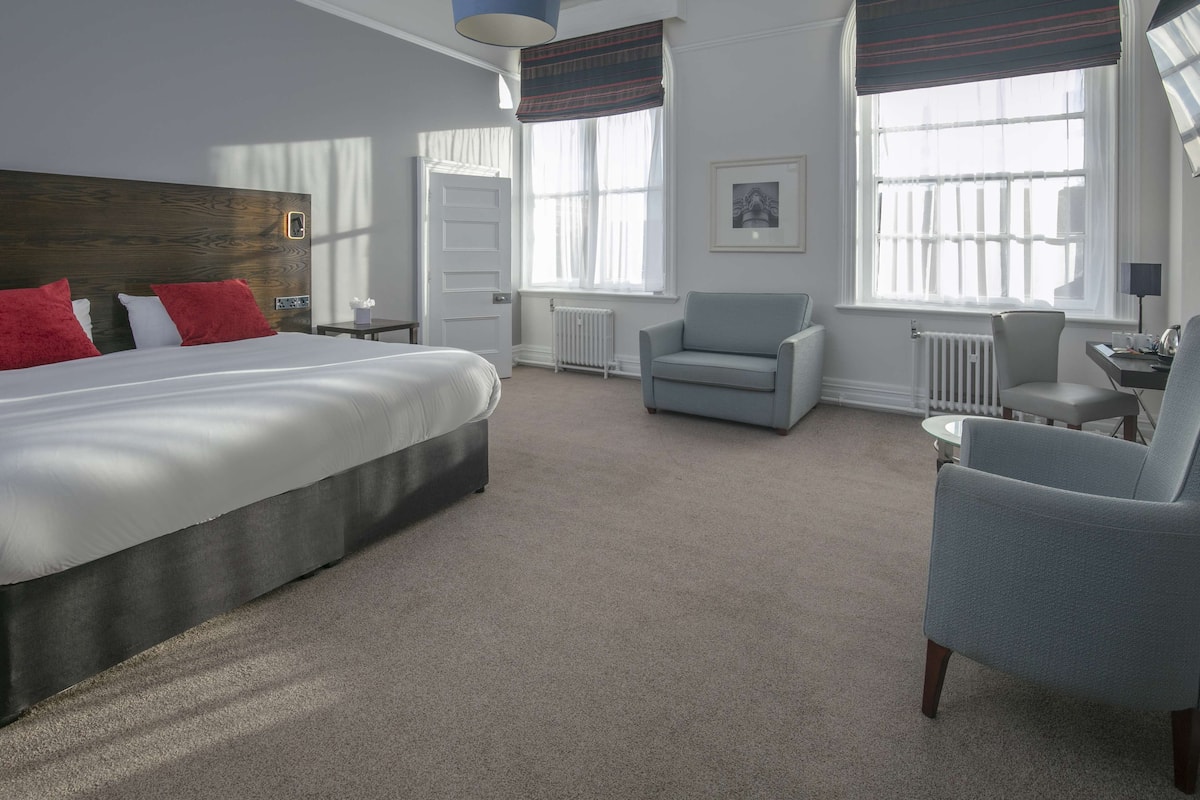 A hotel room with a large bed, two armchairs, a small table, and two windows with curtains.
