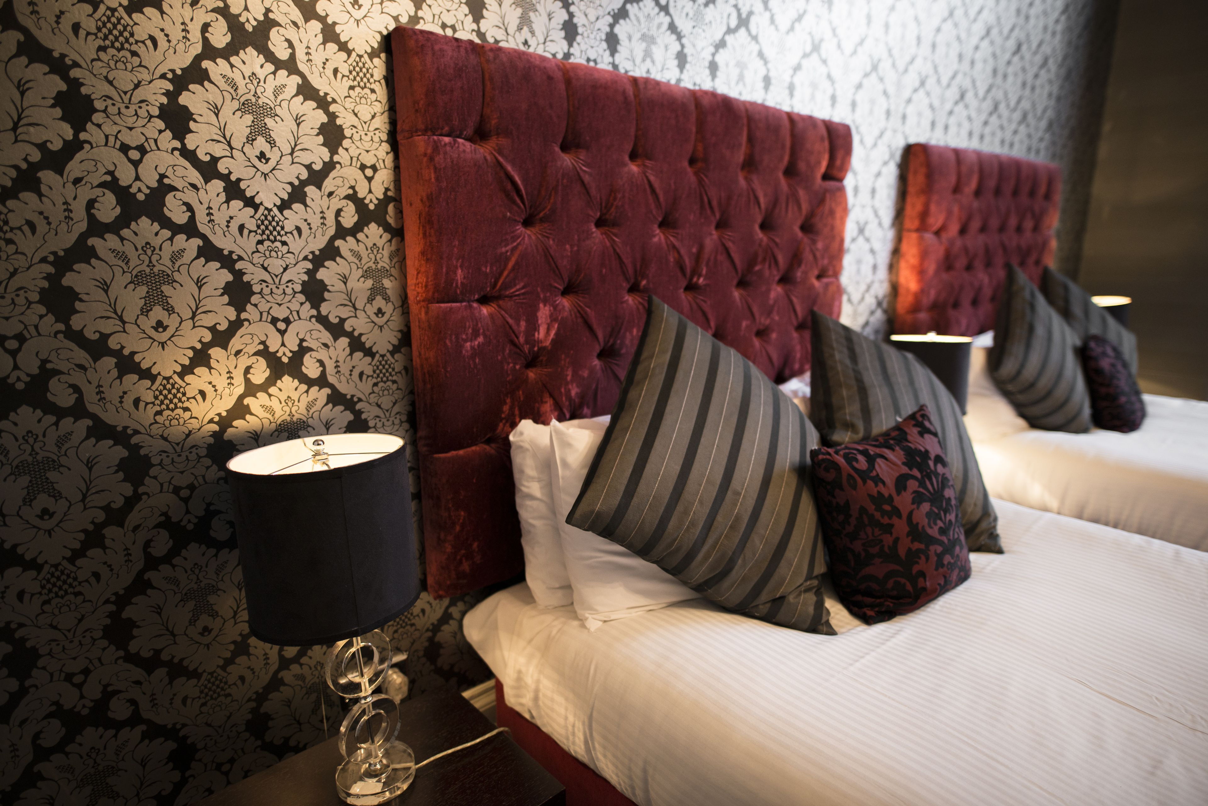 A hotel room with a tufted headboard, a patterned wallpaper, and a bedside lamp.