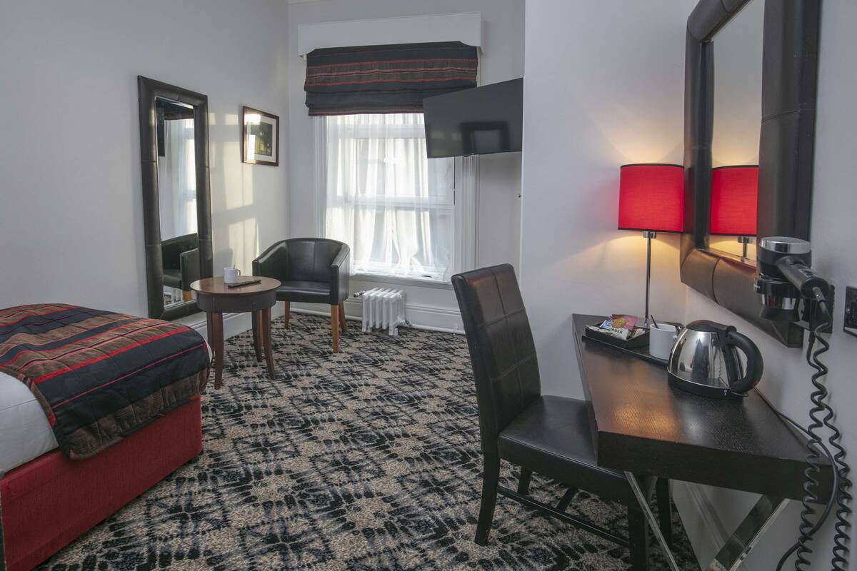 A hotel room with a bed, a desk, a chair, a table, a radiator, a window with curtains, a TV mounted on the wall, and a red lamp.