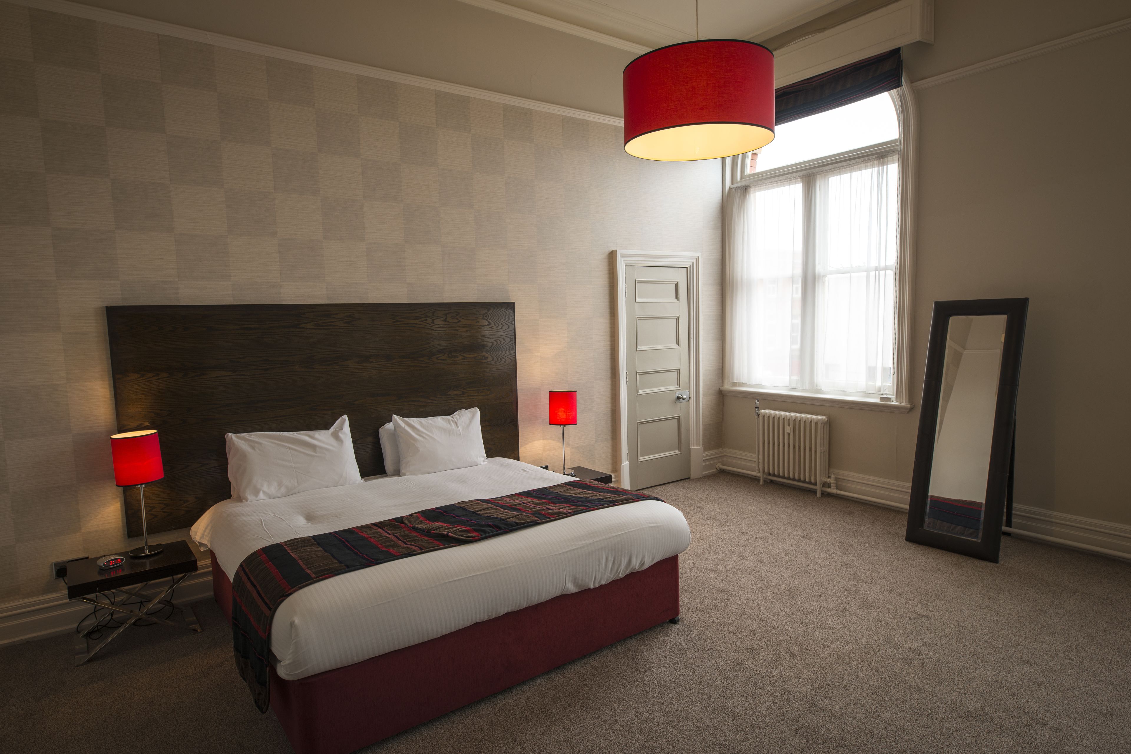 A hotel room with a large bed, a red lamp, a mirror, and a window with curtains.