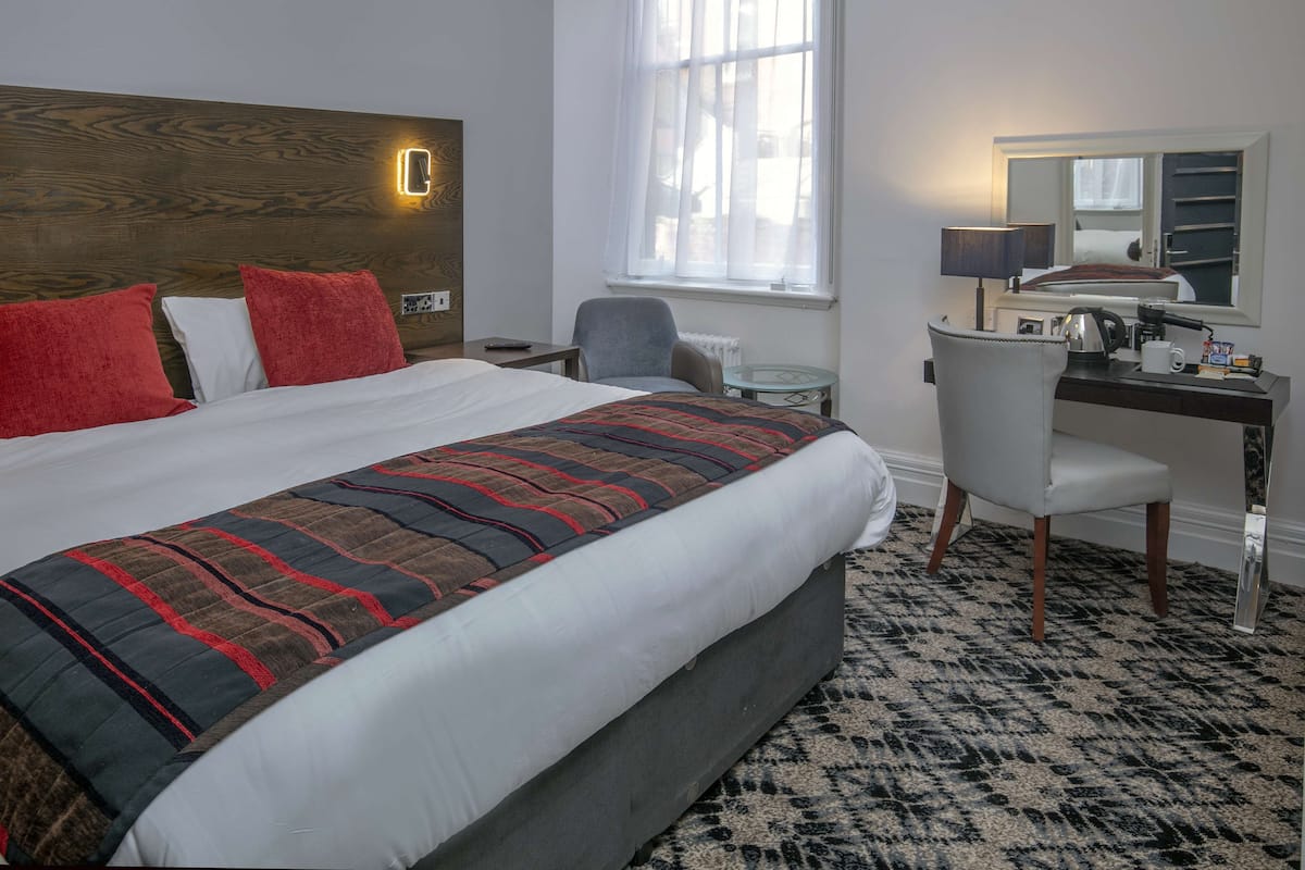 A hotel room with a large bed, a desk with a chair, a mirror, and a window with curtains.