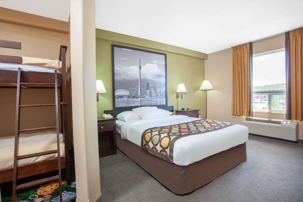 A hotel room with a bunk bed, a large bed with a patterned bedspread, two bedside lamps, a nightstand, a framed cityscape picture on the wall, and a window with curtains.