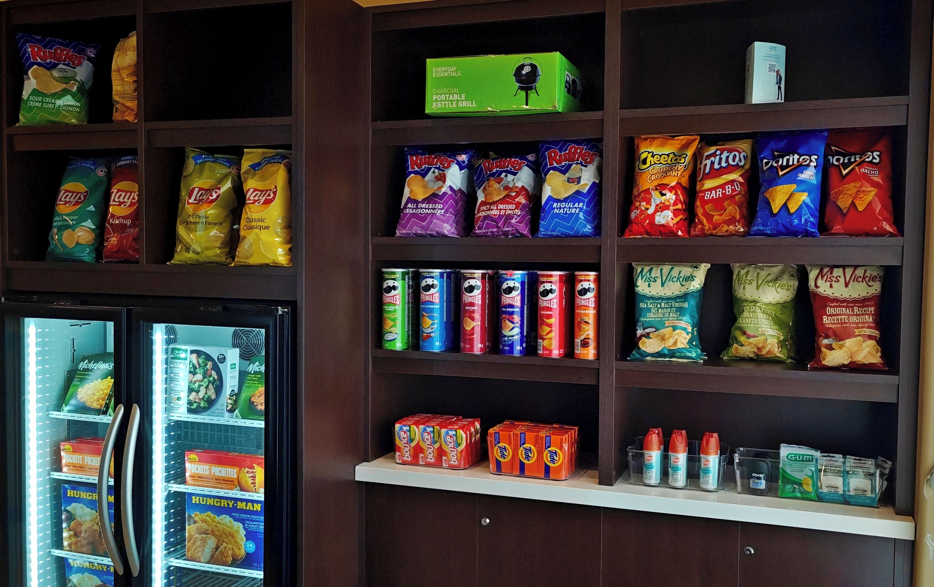 A snack shelf stocked with various chips and snacks, a small refrigerator with frozen foods, and a box of a portable charcoal grill.