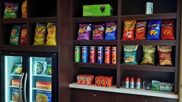A snack shelf stocked with various chips and snacks, a small refrigerator with frozen foods, and a box of a portable charcoal grill.