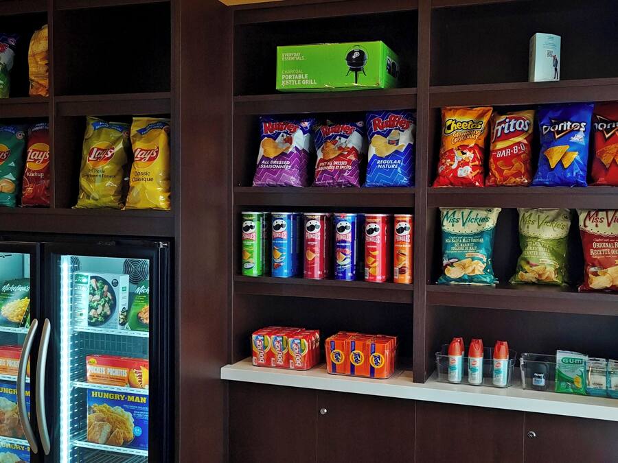 A snack shelf stocked with various chips and snacks, a small refrigerator with frozen foods, and a box of a portable charcoal grill.