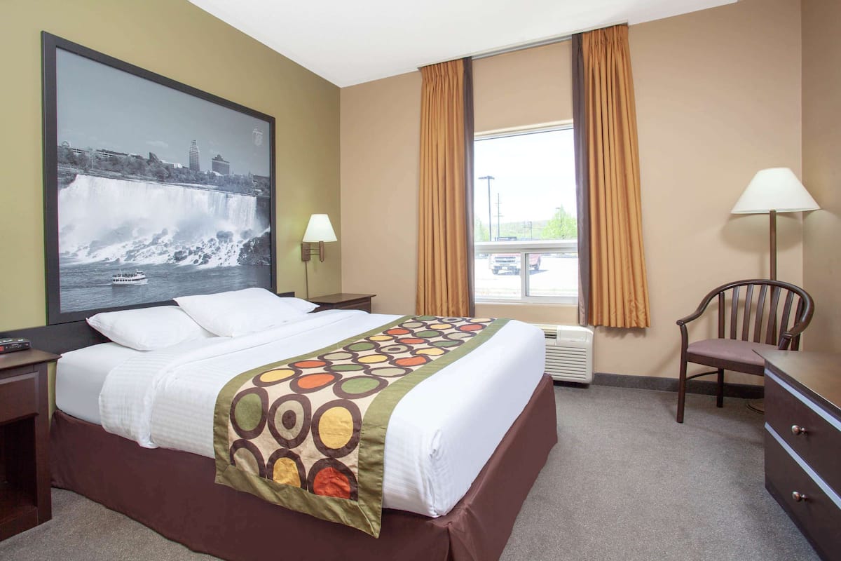 A hotel room with a large bed, a nightstand, a desk, a chair, a lamp, a window with curtains, and a wall-mounted picture of a waterfall.