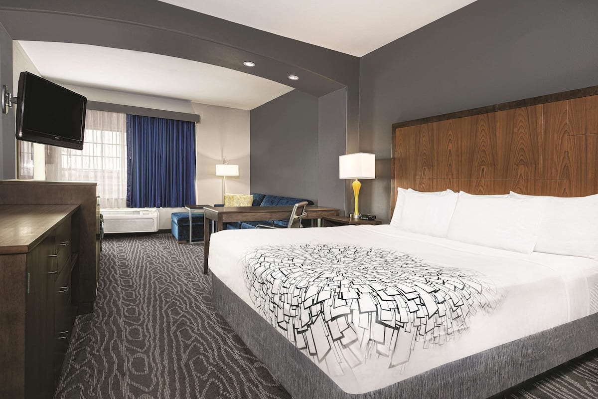 A hotel room with a large bed, a desk with a chair, a flat-screen TV mounted on the wall, and a window with blue curtains.