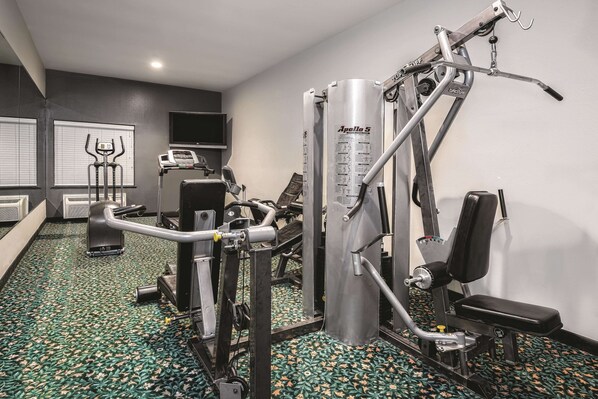Uma academia com diversos equipamentos de exercícios, esteira e televisão fixada na parede.