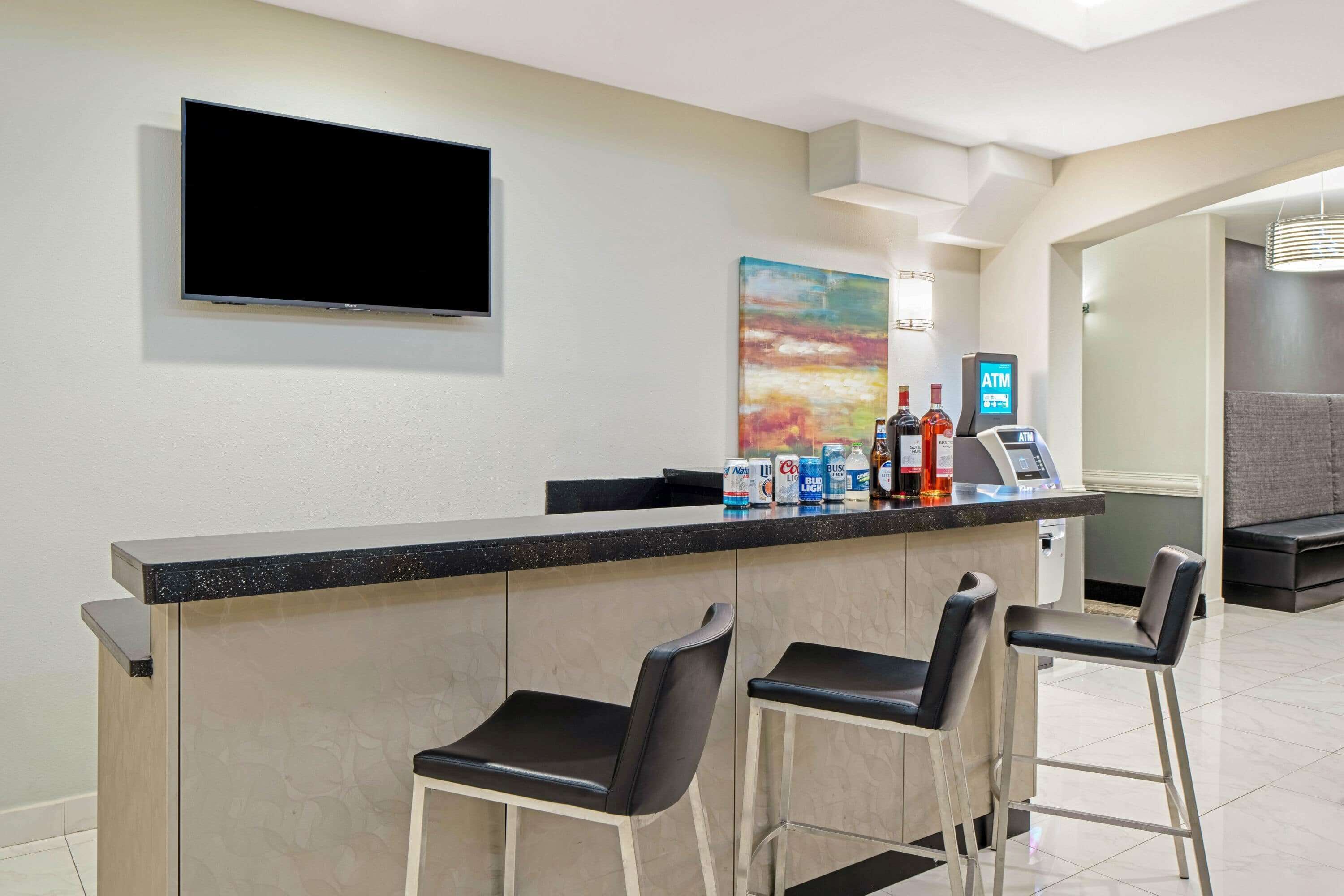 A bar area with a flat-screen TV, a painting, and an ATM. There are three bar stools and a counter with various beverages.