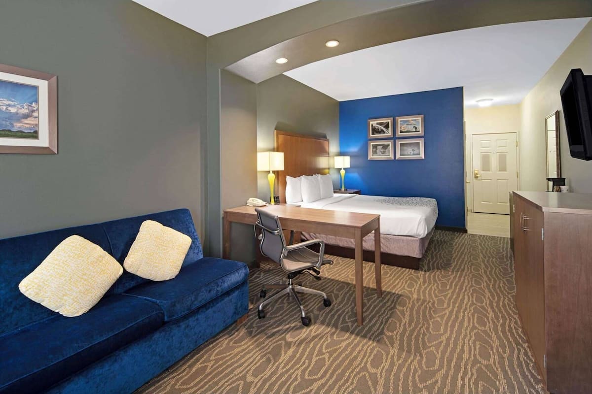 A hotel room with a blue couch, a bed, a desk with a chair, a lamp, and a television.