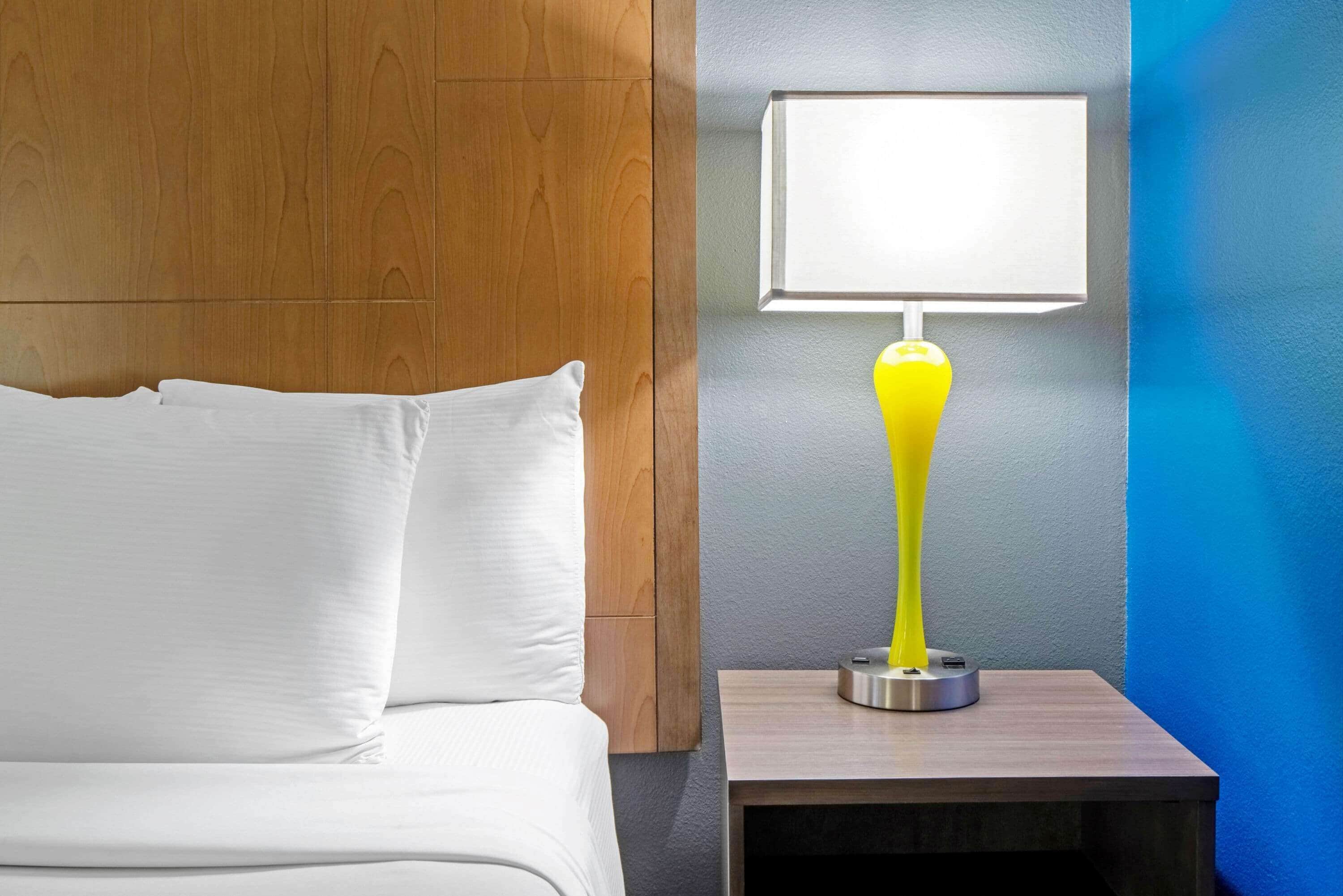 Una cama bien hecha con ropa de cama blanca, cabecera de madera y mesita de noche con una lámpara amarilla moderna.
