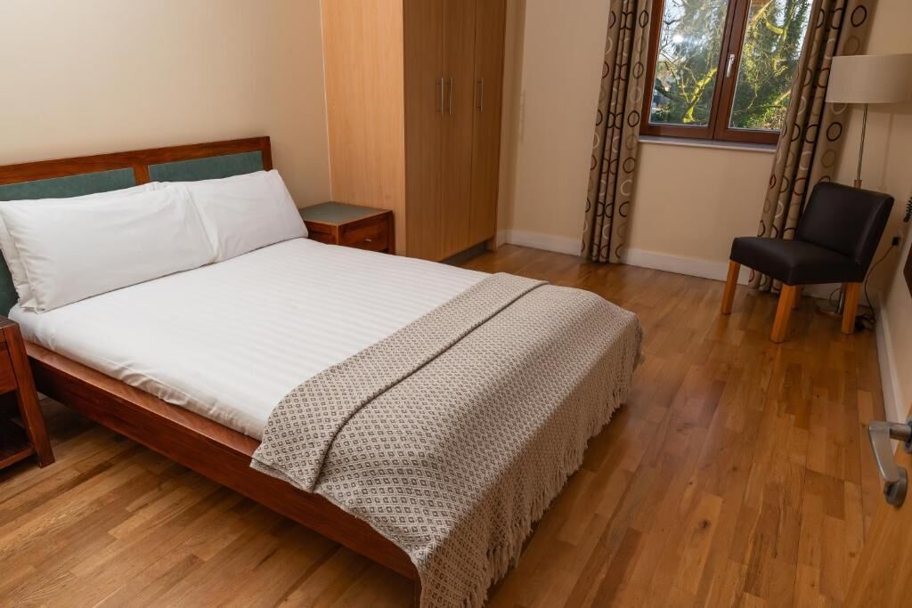 Un dormitorio con cama de madera, mesita de noche, armario, una silla y ventana con cortinas.