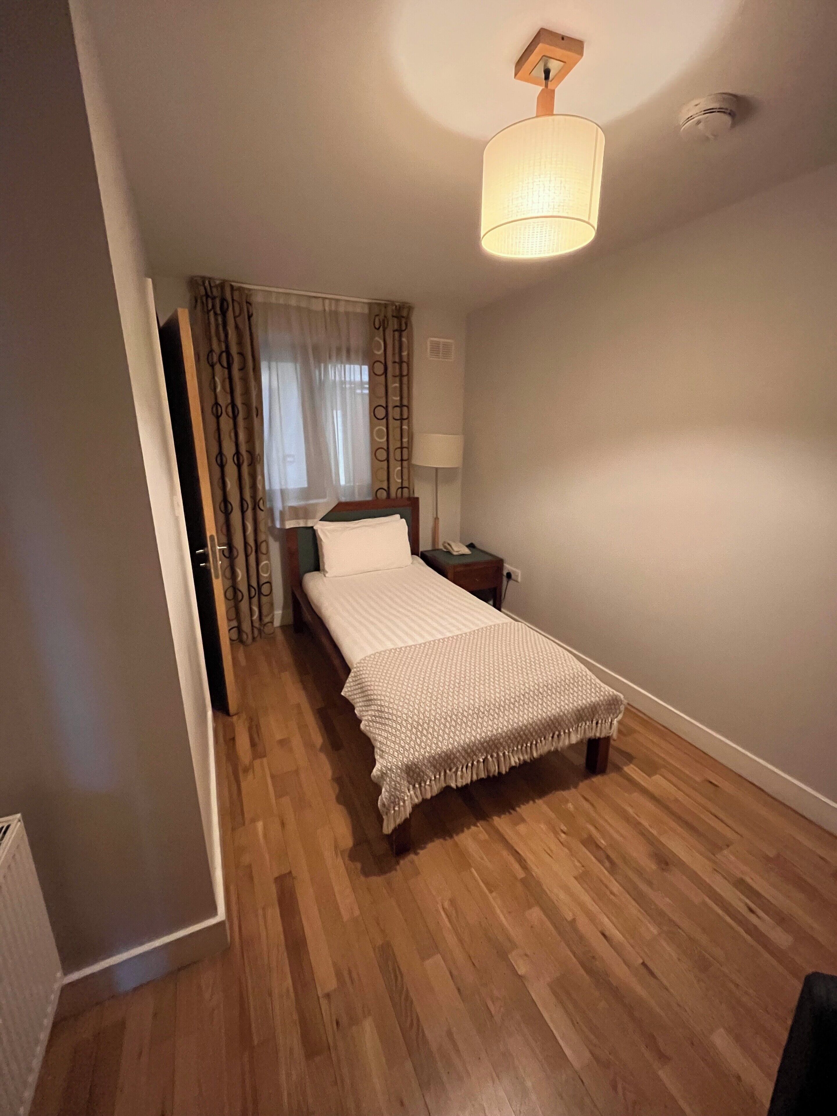 Un dormitorio con una cama individual, una mesita de noche, una lámpara, una luz en el techo y una ventana con cortinas.