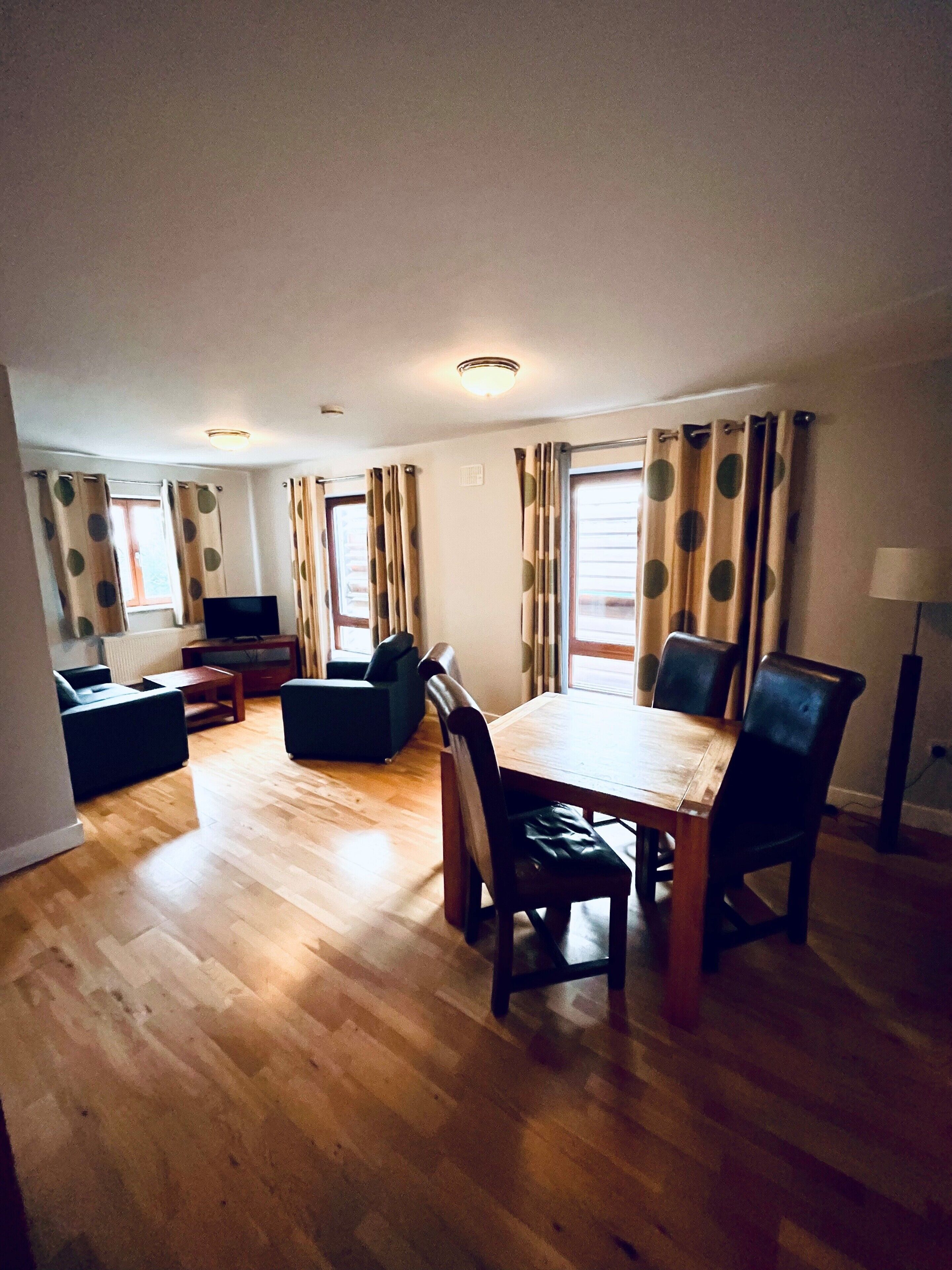 A living room with a wooden floor, a dining table with chairs, a sofa, a TV, and curtains with a polka dot pattern.