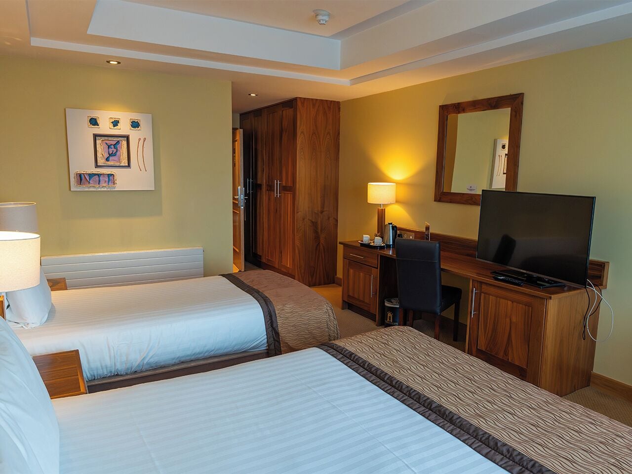 A hotel room with two beds, a desk with a television, a mirror, and a wall-mounted artwork.