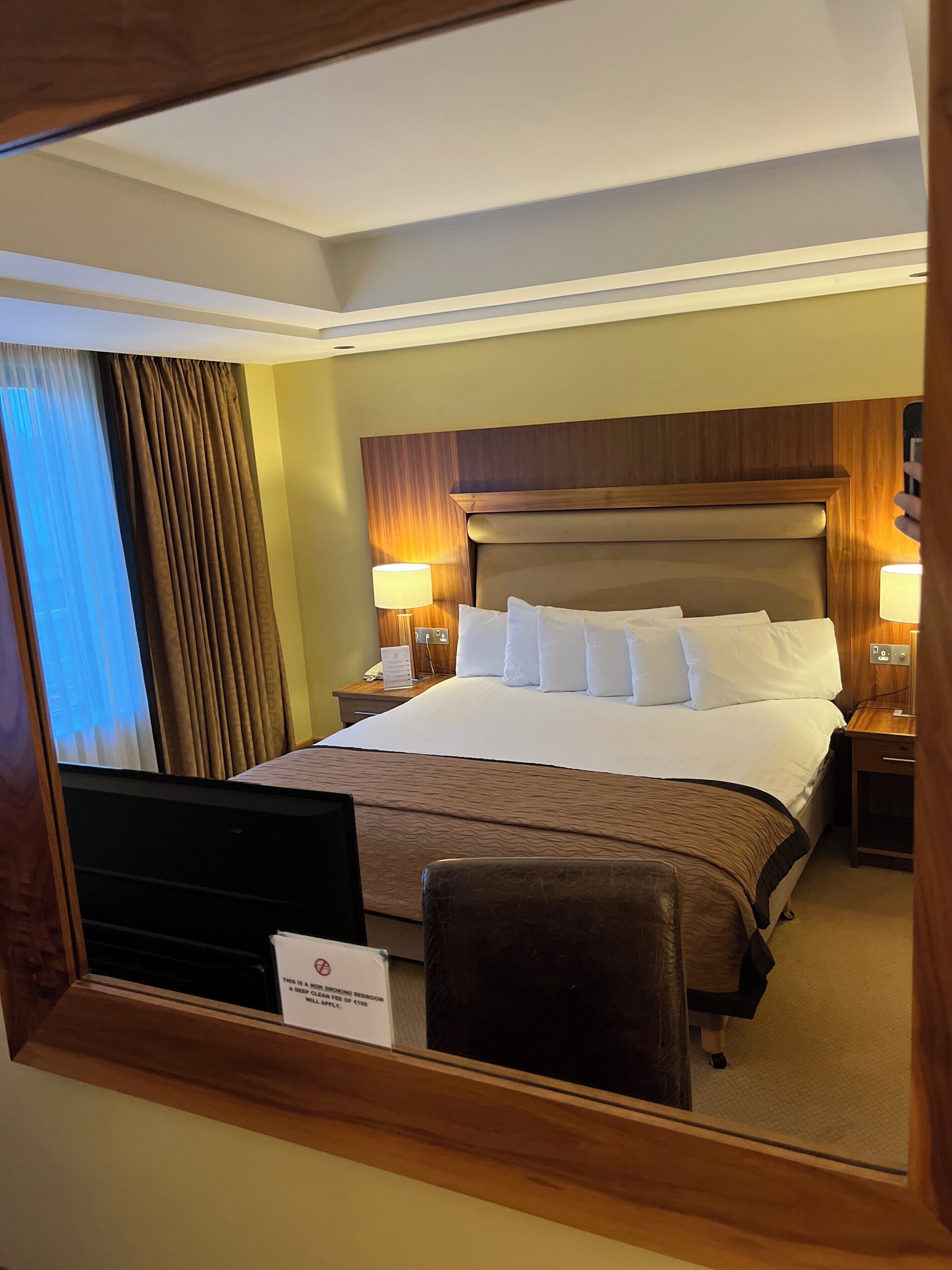 Habitación de hotel con una cama grande, dos mesitas de noche con lámparas, un cabecero de madera y un espejo que refleja la habitación.