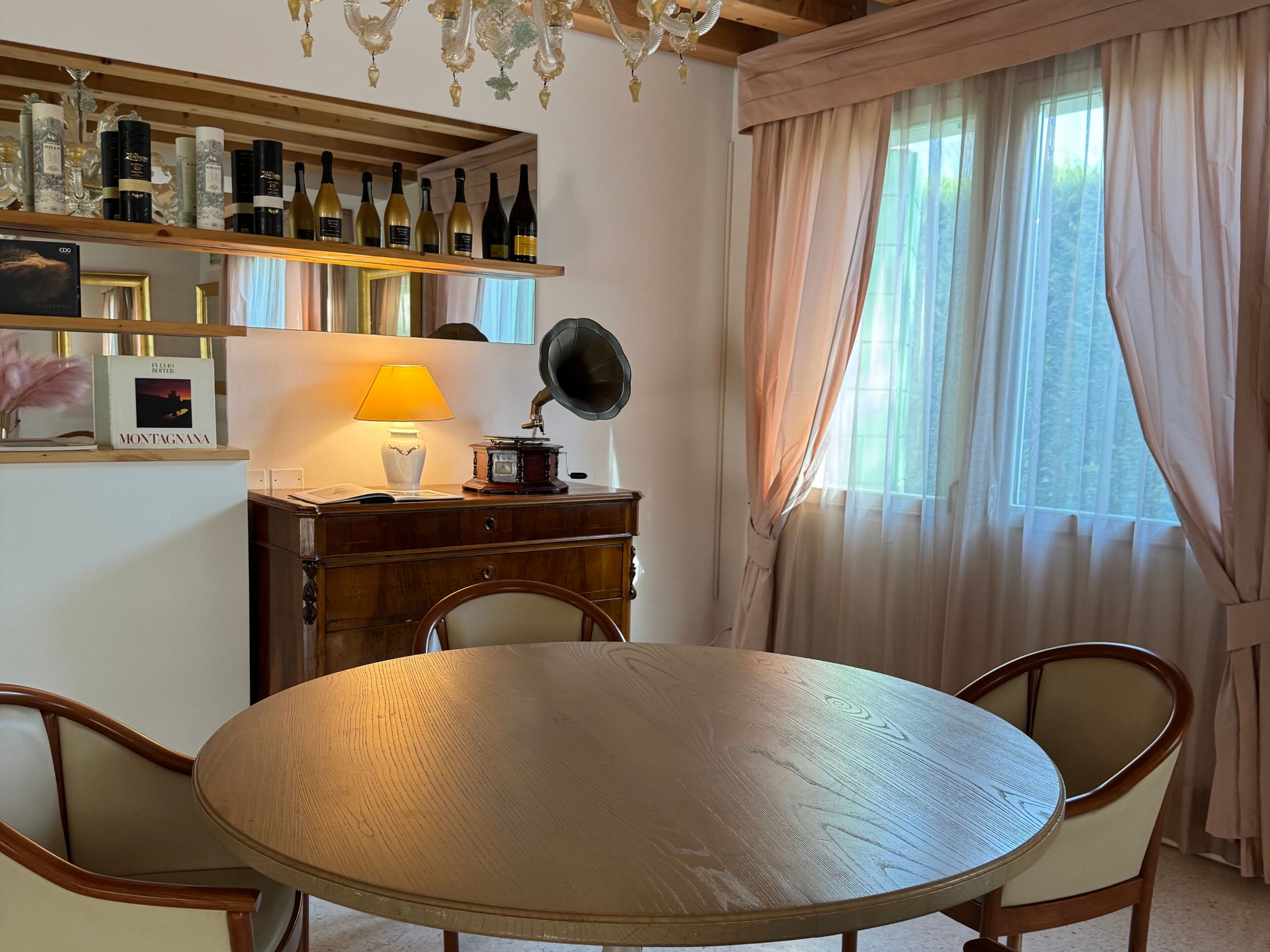 A round wooden table with chairs, a desk with a lamp, a phonograph, and a shelf with wine bottles and a framed picture.