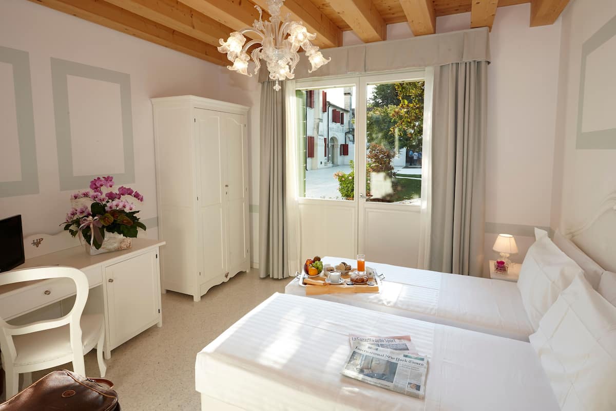 A hotel room with a bed, a desk, a chair, a wardrobe, a chandelier, and a window with curtains.