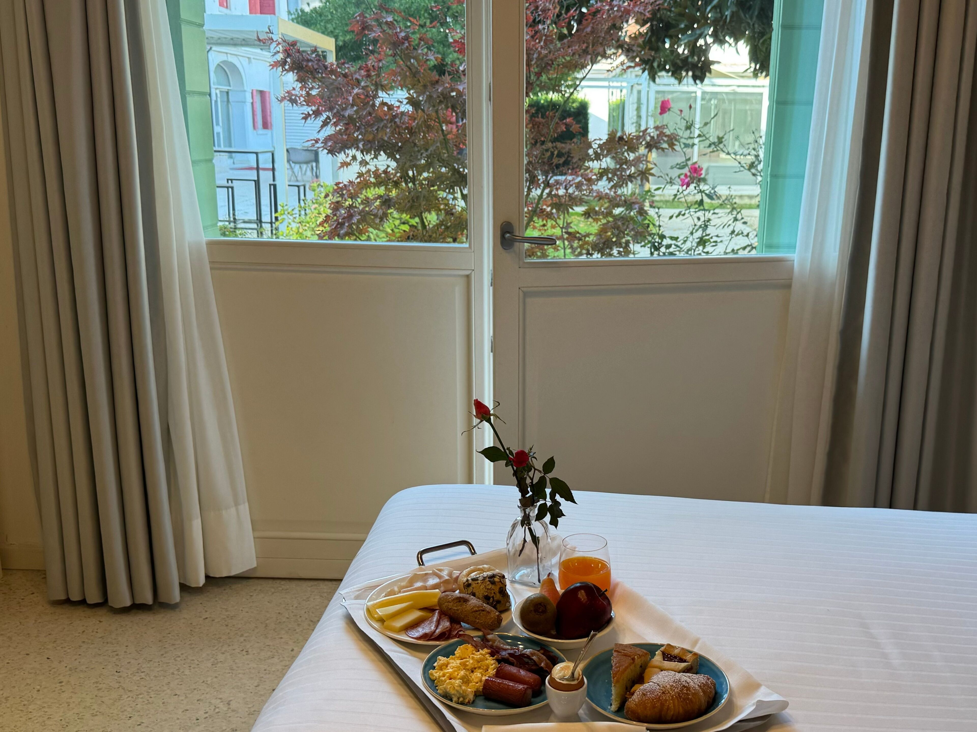 Ein Frühstücksbrett mit verschiedenen Speisen und einem Glas Orangensaft auf einem Bett neben einem Fenster mit Vorhängen.