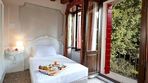 Habitación de hotel con cama, mesita de noche con lámpara, bandeja con alimentos para el desayuno y vistas a zonas verdes a través de una puerta de balcón.