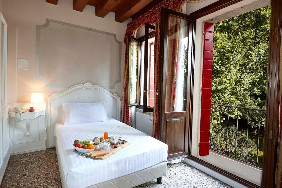 A hotel room with a bed, a bedside table with a lamp, a tray with breakfast items, and a view of greenery through a balcony door.