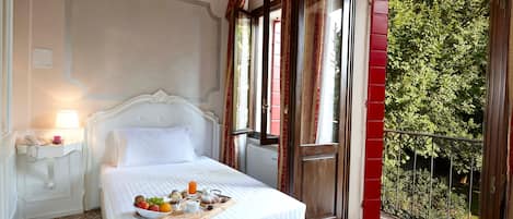A hotel room with a bed, a bedside table with a lamp, a tray with breakfast items, and a view of greenery through a balcony door.