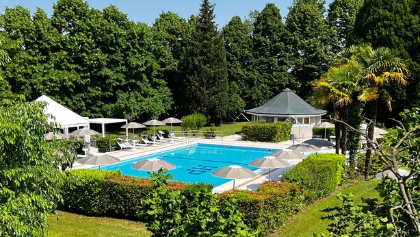 Una piscina rodeada de sillas de descanso y sombrillas, con un pabellón y árboles frondosos al fondo.