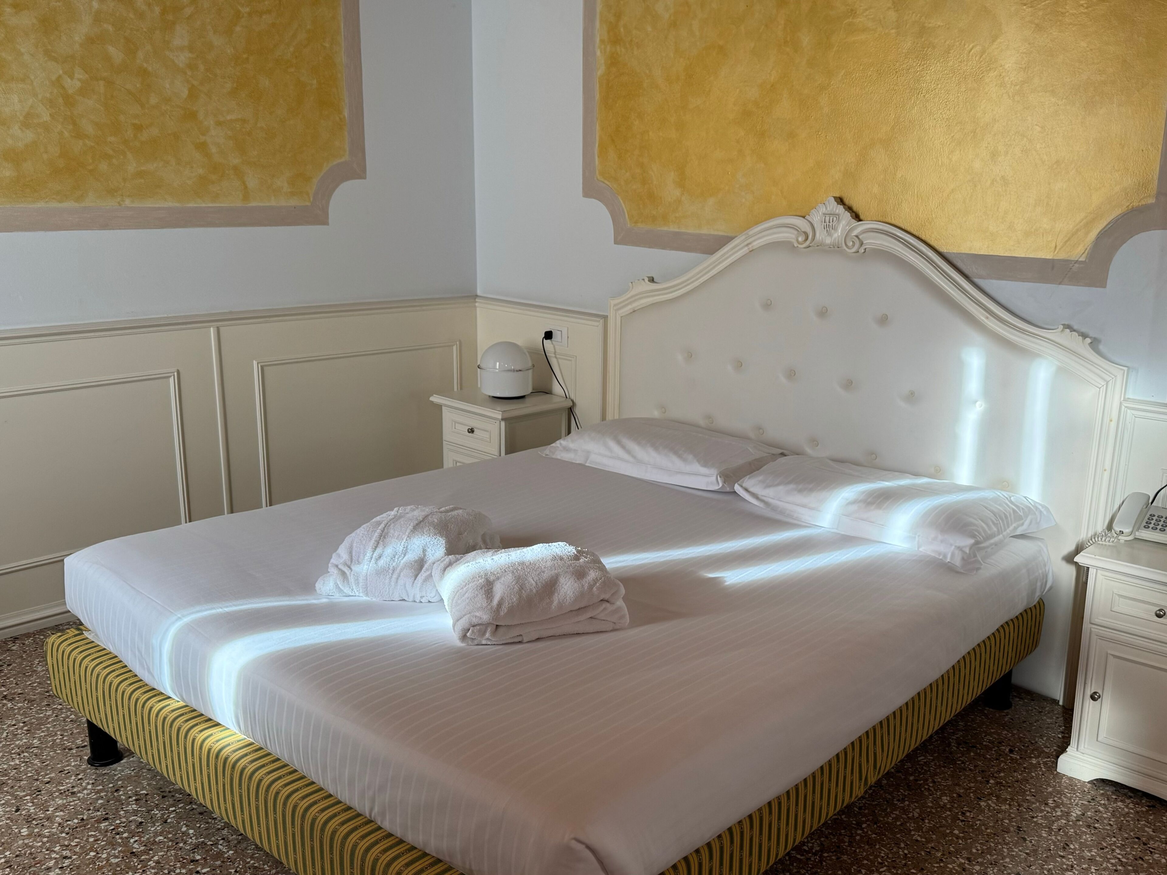 Una cama bien hecha con sábanas blancas y dos toallas, una mesita de noche con lámpara, y una pared con detalles dorados.