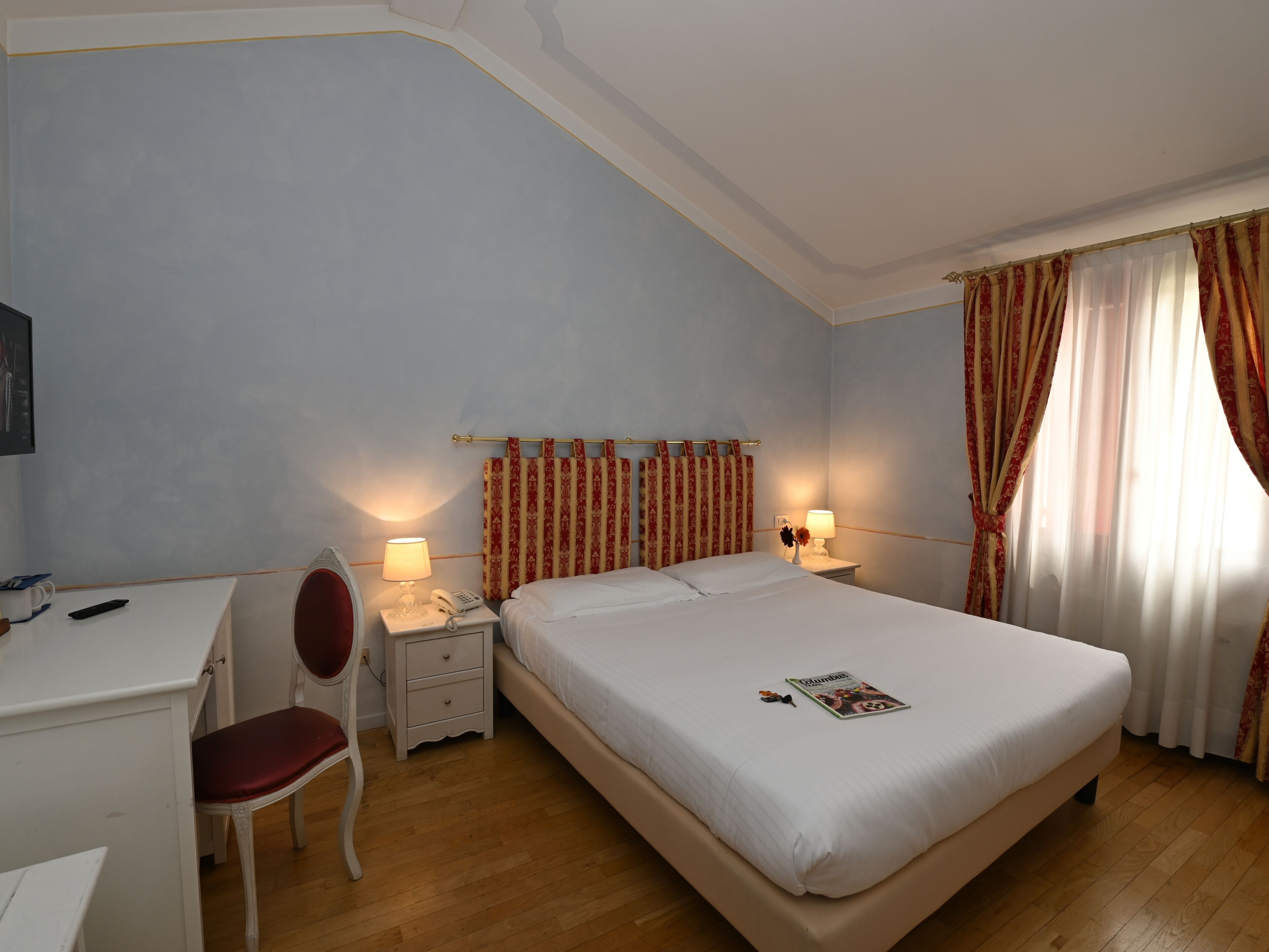 A hotel room with a bed, a desk, a chair, a nightstand with a lamp, and a window with curtains.