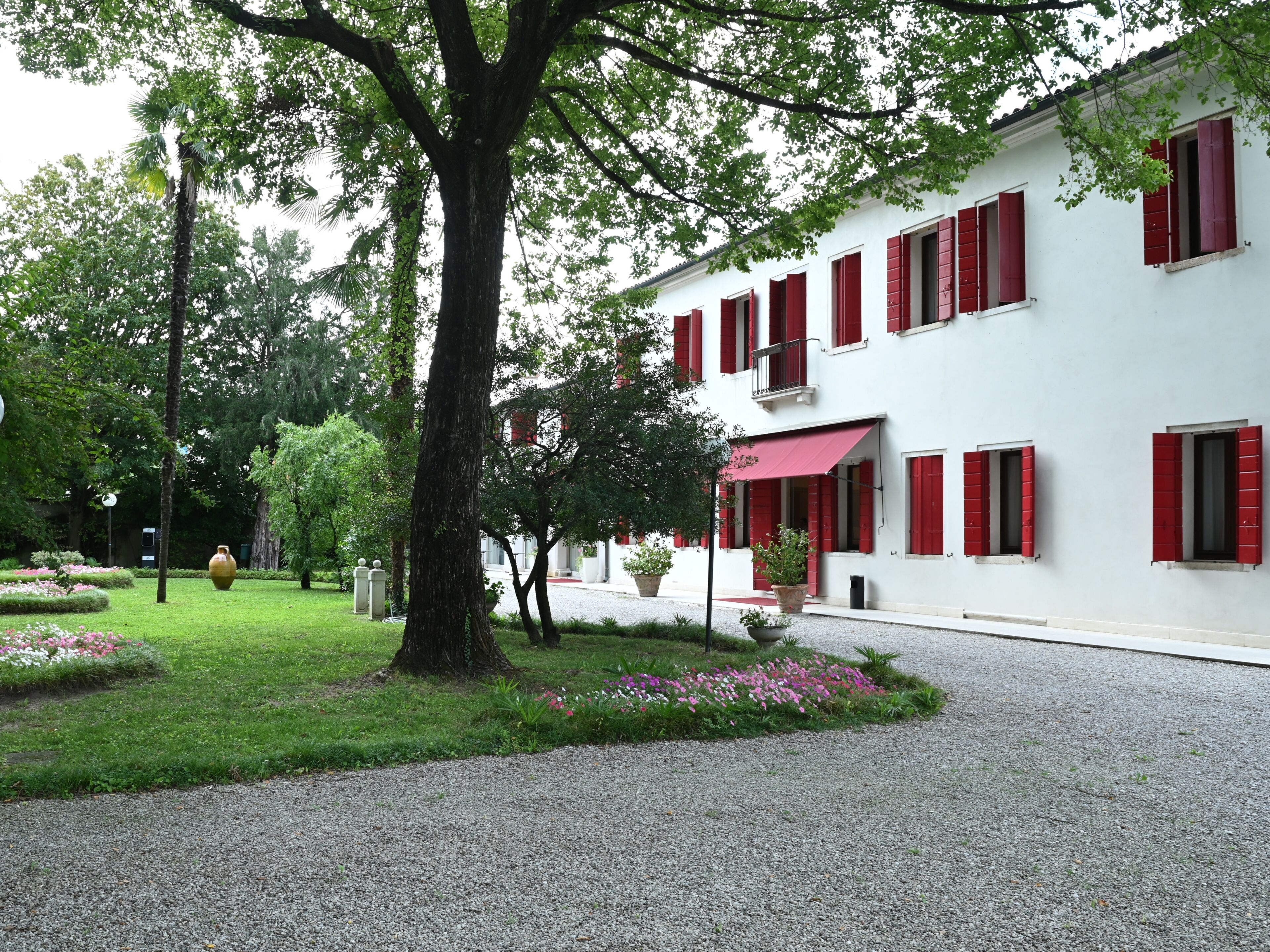 Un bâtiment aux volets rouges, une allée gravillonnée et un jardin bien entretenu agrémenté de fleurs roses.