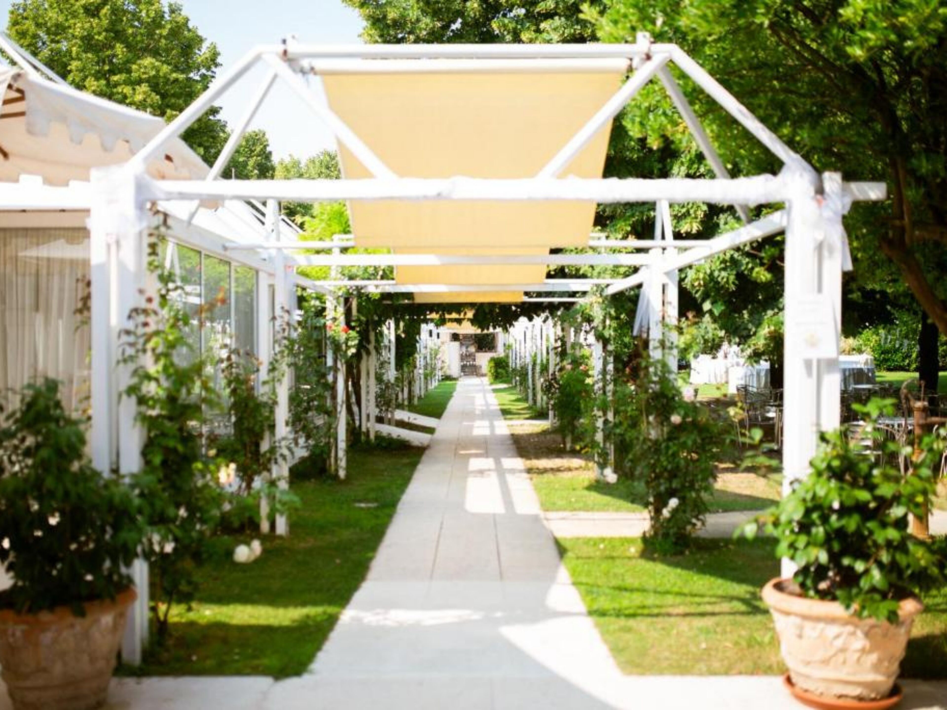A pathway lined with white arches and potted plants, leading to a covered area with a beige canopy.