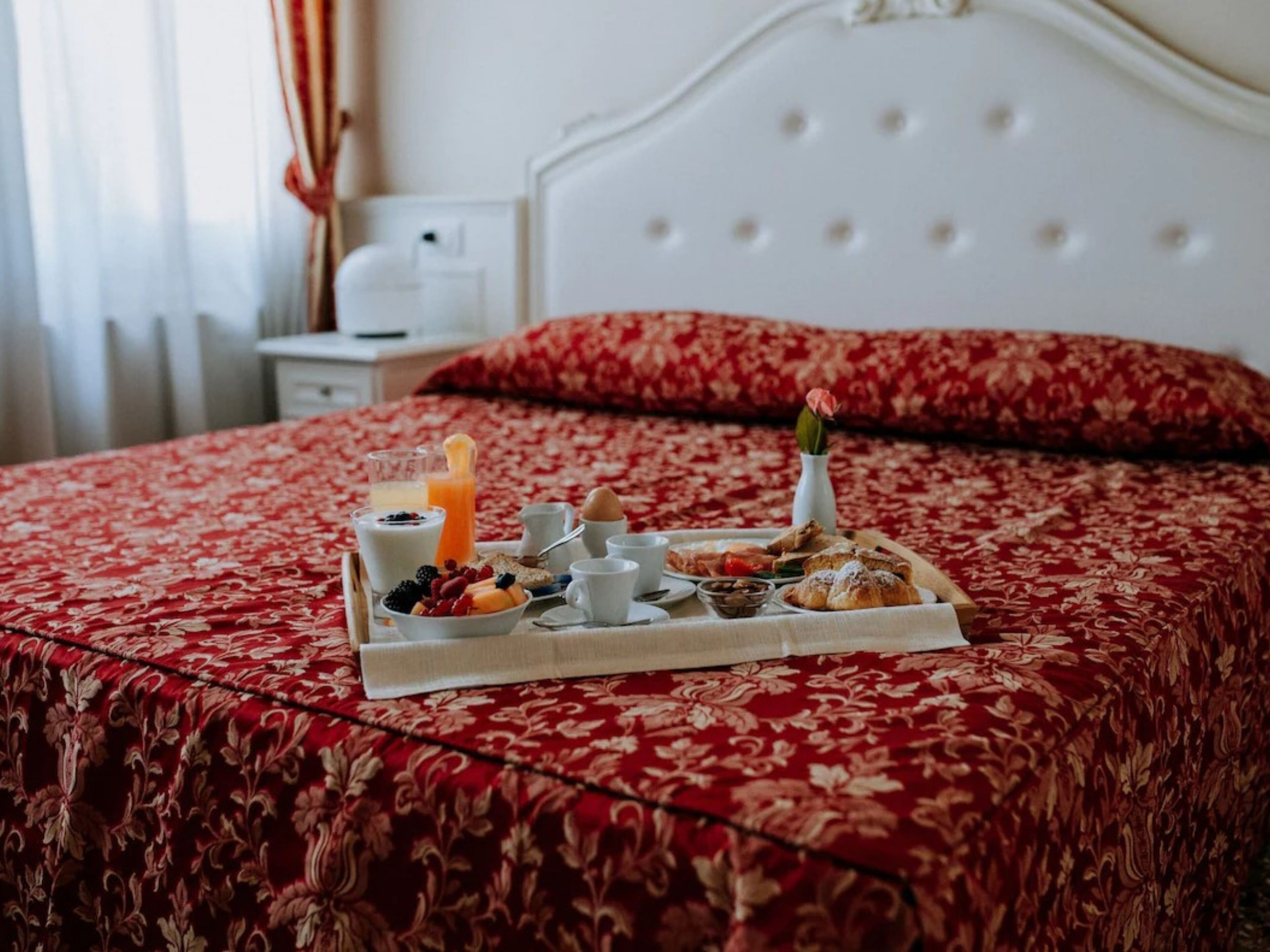 Un plateau de petit-déjeuner soigneusement préparé est posé sur un lit recouvert d’une courtepointe à motifs floraux rouges, avec une tête de lit blanche et un oreiller rouge.
