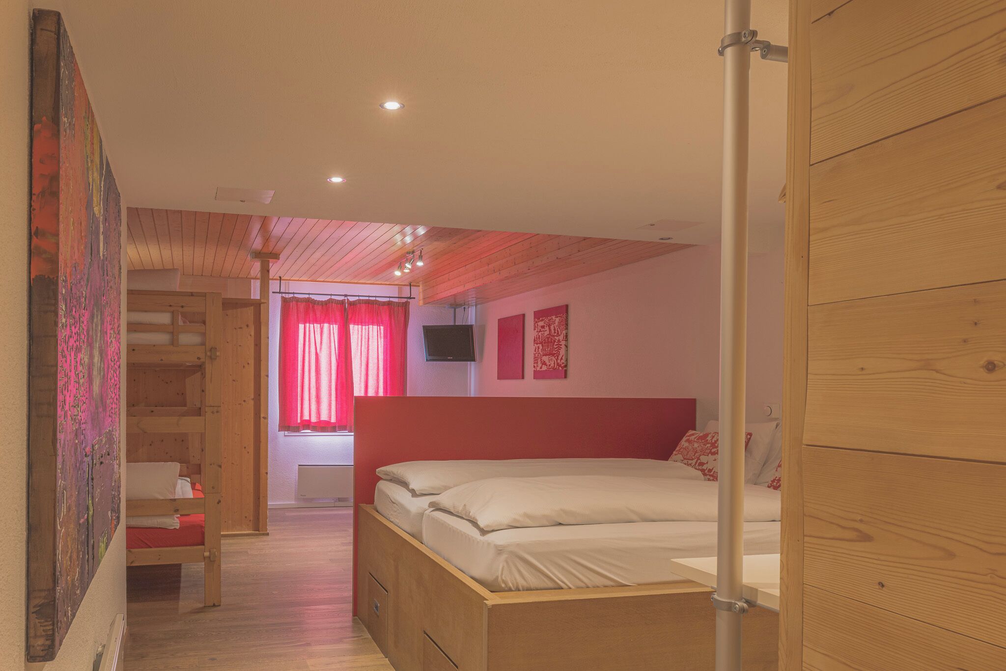 Una habitación con literas, un escritorio y una silla. Se ve una ventana con cortinas rojas y un televisor en la pared.