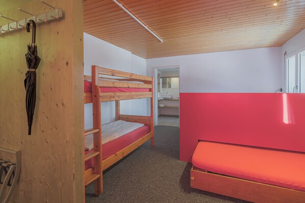Habitación con literas, colchón rojo, techo de madera y un baño pequeño visible a través de una puerta abierta.