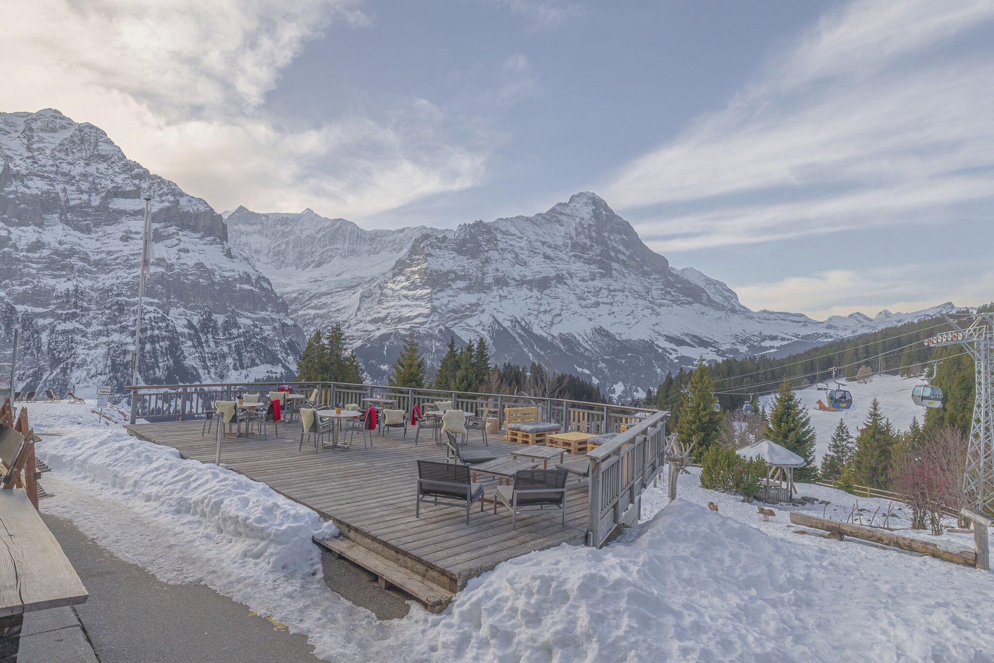 Una terraza de madera con mobiliario exterior, con vistas a un paisaje montañoso nevado.