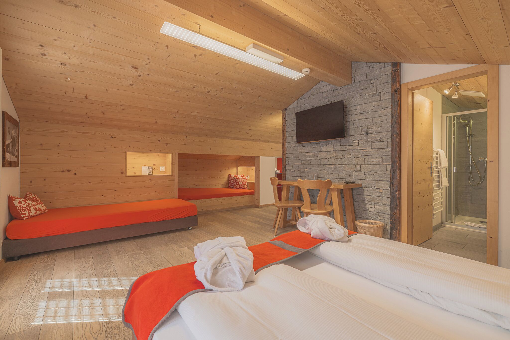 A modern hotel room with a wooden ceiling, a stone wall, a flat-screen TV, a shower, and a bed with a red blanket.