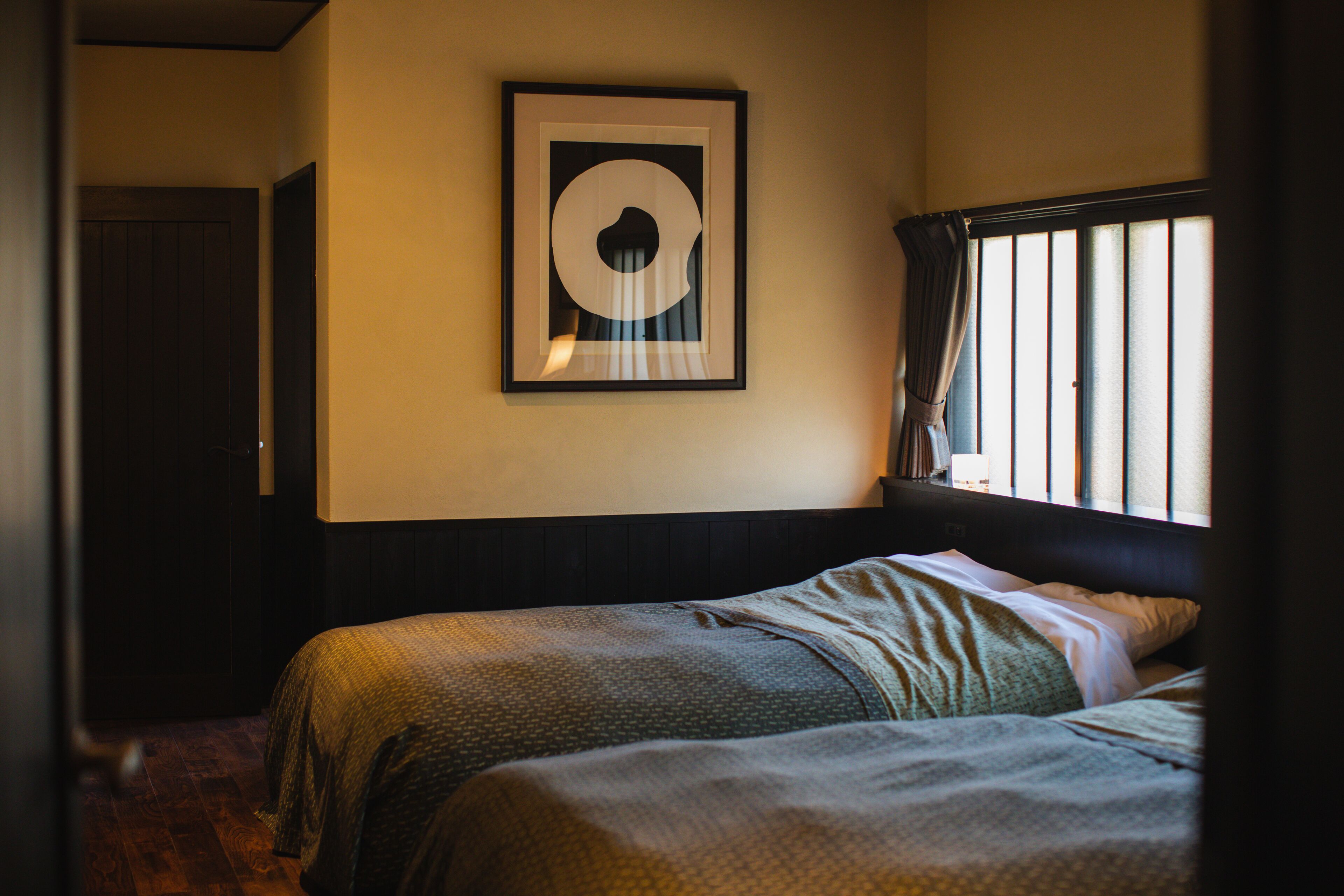A bedroom with two beds, a framed artwork on the wall, and a window with blinds.