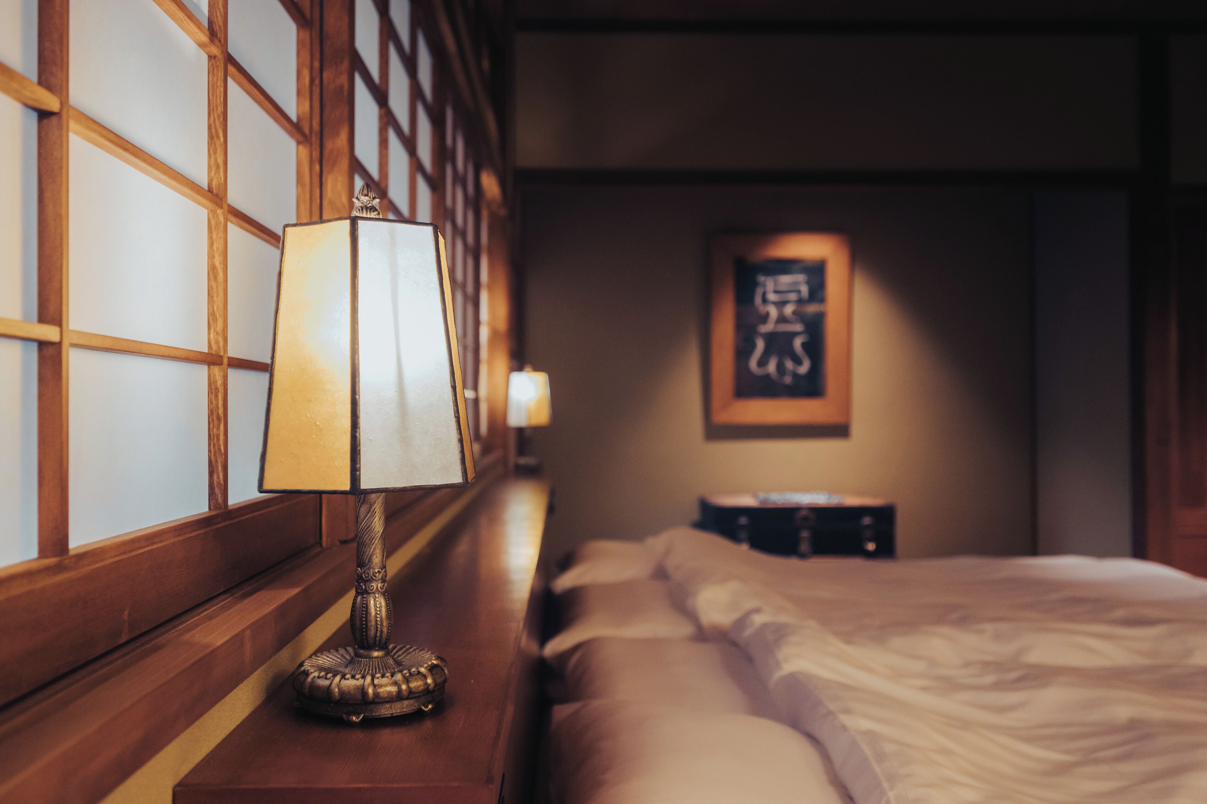 A hotel room with a wooden headboard, a bedside lamp, a bed with white linens, and a framed artwork on the wall.